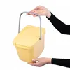 Hands holding a yellow plastic waste bin with a lid and a gray handle. Produktbild-Vorschau