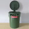 A green trash can with its lid open, placed on a wooden surface against a textured wall. Produktbild-Vorschau