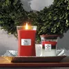 Two WoodWick candles are displayed on a decorative tray in front of a green wreath. Podgląd zdjęcia produktu
