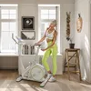 A woman is standing next to a white exercise bike in a bright, modern room with large windows. Produktbild-Vorschau