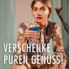 A woman holds a blue cocktail with a fig garnish, looking at the camera. She is wearing a floral shirt and a grey apron-like garment. The background is a soft green curtain and a beige wall. Produktbild-Vorschau