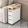 A narrow white cabinet with wooden accents and drawers is placed next to a sofa. The top surface of the cabinet holds a magazine, a cup, and a small wooden tray. Produktbild-Vorschau