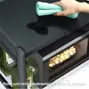 A hand is wiping a wet surface on a black kitchen shelf, with a microwave and bottles of olive oil visible below. Náhľad obrázku produktu