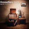 A vintage-style music player with a turntable, radio, and cassette deck, set on a wooden stand next to a plant and a lamp. A cassette tape is mounted on the wall above. Produktbild-Vorschau