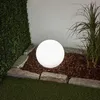 A glowing spherical garden light sits in a garden bed with mulch and stones, next to a hedge and tall grass. Náhľad obrázku produktu