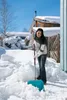 A woman is shoveling snow in a yard with houses and trees in the background. Náhled obrázku produktu