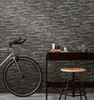 A bicycle is parked next to a dark wooden desk with a stool in front of it, against a stone-patterned wall. Náhľad obrázku produktu