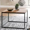 A square coffee table with a wooden top and black metal legs, placed on a patterned rug in a living room setting. Decorative items like a bowl of snacks, a plant, and a candle are on the table. Produktbild-Vorschau