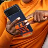 A person wearing an orange sweater is holding a blue phone case with an orange strap. Produktbild-Vorschau