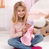 A young girl is sitting on the floor, holding a baby doll. The doll is wearing a pink outfit and a gray and pink hat. The girl is smiling at the camera. Produktbild-Vorschau