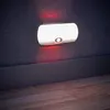 A white LED night light with a motion sensor is mounted on a dark textured wall above a dark gray baseboard. The floor in front of the baseboard is made of light brown wood planks. Produktbild-Vorschau