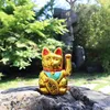 A golden Maneki Neko cat figurine sits on a rock in a garden setting. Produktbild-Vorschau