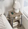 A small wooden side table with two shelves next to a bed, featuring a lamp and a phone charging on top. Produktbild-Vorschau