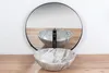 A marble sink with a black faucet is placed in front of a large round mirror, reflecting the sink and faucet. The scene is set against a white wall and a wooden floor. Podgląd zdjęcia produktu