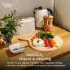 A cheese board with various food items arranged on a wooden table, with kitchen appliances in the background. Produktbild-Vorschau