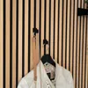 Two black hooks are mounted on a slatted wooden wall. One hook holds a tan handbag with a chain strap, and the other holds a black hanger with a light-colored jacket on it. Produktbild-Vorschau