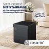 A black storage ottoman is placed on a textured rug in a living room setting, with a side table and flowers in the background. Produktbild-Vorschau