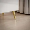 Close-up shot of the bottom of a white cabinet with gold legs on a wooden floor. Produktbild-Vorschau