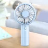 A light blue handheld fan is placed on a wooden table in front of a blurred background of a living room with a person lying on a couch. Produktbild-Vorschau