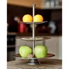 A three-tiered serving stand with fruit and a nut on a wooden table, with a kitchen in the background. Produktbild-Vorschau