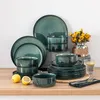 A collection of teal-colored ceramic dishes, including plates and bowls, arranged on a wooden surface with lemons and a vase of yellow flowers in the background. Náhled obrázku produktu