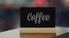 A small black chalkboard with the word "Coffee" written on it in chalk, resting on a wooden stand on a table. Produktbild-Vorschau