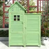 A green wooden garden shed with a small window and two doors, set against a blurred background of foliage and a wooden trellis. Náhled obrázku produktu