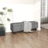 A dark gray storage bench with a black and white checkered blanket draped over it, placed in a room with wooden flooring and a large plant in a glass planter. Produktbild-Vorschau