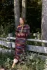 A woman leans against a wooden fence in a wooded area, wearing a striped dress and boots. Produktbild-Vorschau