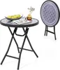 A round folding table with a patterned top, set with a bowl of apples, a glass, and a vase with dried flowers. A second table is shown folded in the background.