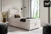 A bedroom scene featuring a single bed with a headboard, pillows, and a throw blanket. A bedside table with a vase of dried flowers is to the left, and a window with curtains is to the right. A black wall lamp is mounted above the bed. Produktbild-Vorschau