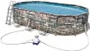 An oval above-ground swimming pool with a stone-like pattern on its sides, connected to a pump and filter system. Náhled obrázku produktu