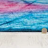 A close-up view of a rug with abstract blue and pink lines, placed on a light-colored wooden surface. A coin is visible on the right side, indicating scale. Náhled obrázku produktu