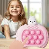 A young girl is smiling and looking at the camera while sitting at a table with a pink electronic game in front of her. A finger is shown pressing one of the buttons on the game. Produktbild-Vorschau