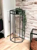 A black metal plant stand with a trailing plant on it, placed on a wooden floor next to a white door and a wood-paneled wall. Produktbild-Vorschau