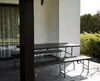 A picnic table and two benches are set up on a patio with a white wall and green bushes in the background. Podgląd zdjęcia produktu