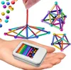 A hand holding a tin of magnetic balls and rods, with several magnetic structures built from them in the background. Náhled obrázku produktu