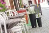 An outdoor cafe setting with tables and chairs, and two large planters with plants. Produktbild-Vorschau