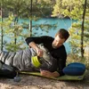 A man is lying in a sleeping bag outdoors by a lake, pouring liquid from a collapsible container into a cup. Produktbild-Vorschau