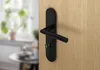 A black door handle set on a wooden door, with a blurred background of a room. Produktbild-Vorschau