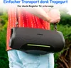 A person is holding a portable speaker with a strap over their shoulder, outdoors. Produktbild-Vorschau