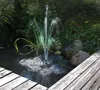 A solar-powered fountain pump operating in a pond surrounded by greenery and rocks, with a wooden walkway in the foreground. Produktbild-Vorschau
