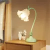 A flower-shaped desk lamp sits on a wooden nightstand next to a bed. Produktbild-Vorschau