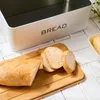A white bread box with the word "BREAD" printed on it, sits on a white tiled surface. Next to it, on a wooden cutting board, are slices of bread and a loaf of bread. A small dish of butter is also visible. Podgląd zdjęcia produktu