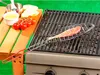 A fish grilling basket with a fish inside is placed on a grill, with some ingredients like tomatoes and bread on a wooden surface next to it. Náhled obrázku produktu