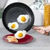 A hand is holding a frying pan with fried eggs in it, and a plate with toast and eggs is below. Produktbild-Vorschau