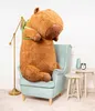 A giant plush capybara sits in a light blue armchair next to a floor lamp and small tables with decorative items. Náhled obrázku produktu