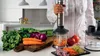 A person is using an immersion blender to chop vegetables in a kitchen setting. Anteprima dell’immagine prodotto