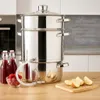 A stainless steel steam juicer is set up on a wooden countertop in a kitchen setting. There are bottles of juice, a bowl of raspberries, and apple slices nearby. Produktbild-Vorschau