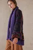 A woman wearing a purple cardigan with a patterned sleeve, standing against a concrete wall. Produktbild-Vorschau